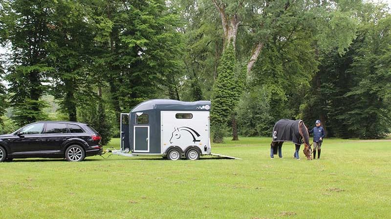 Un furgone agganciato a un'auto in un prato verde. Un cavallo e un cavaliere pascolano nelle vicinanze.