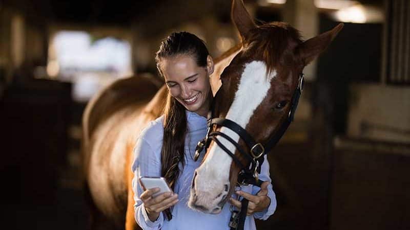 Giovane donna che guarda il suo smartphone e sorride. Il suo cavallo si guarda alle spalle.